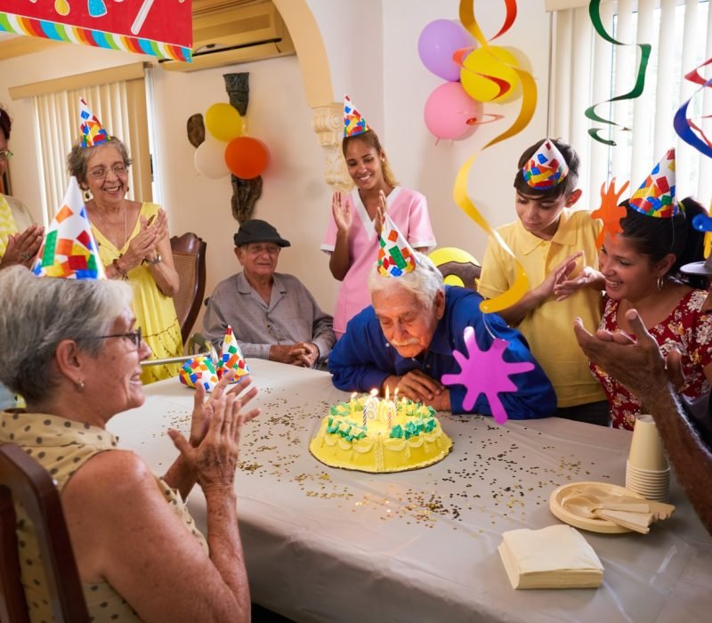 Group of old friends and family celebrating senior man 80 birthday party in retirement home. Happy elderly people having fun. Grandfather blowing candles on cake.